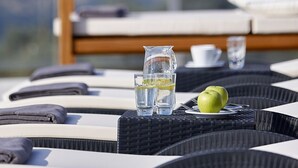 Un espacio al aire libre iluminado por el sol con una mesa para dos, que incluye una jarra de vidrio transparente, dos vasos, un plato con dos manzanas y una taza.