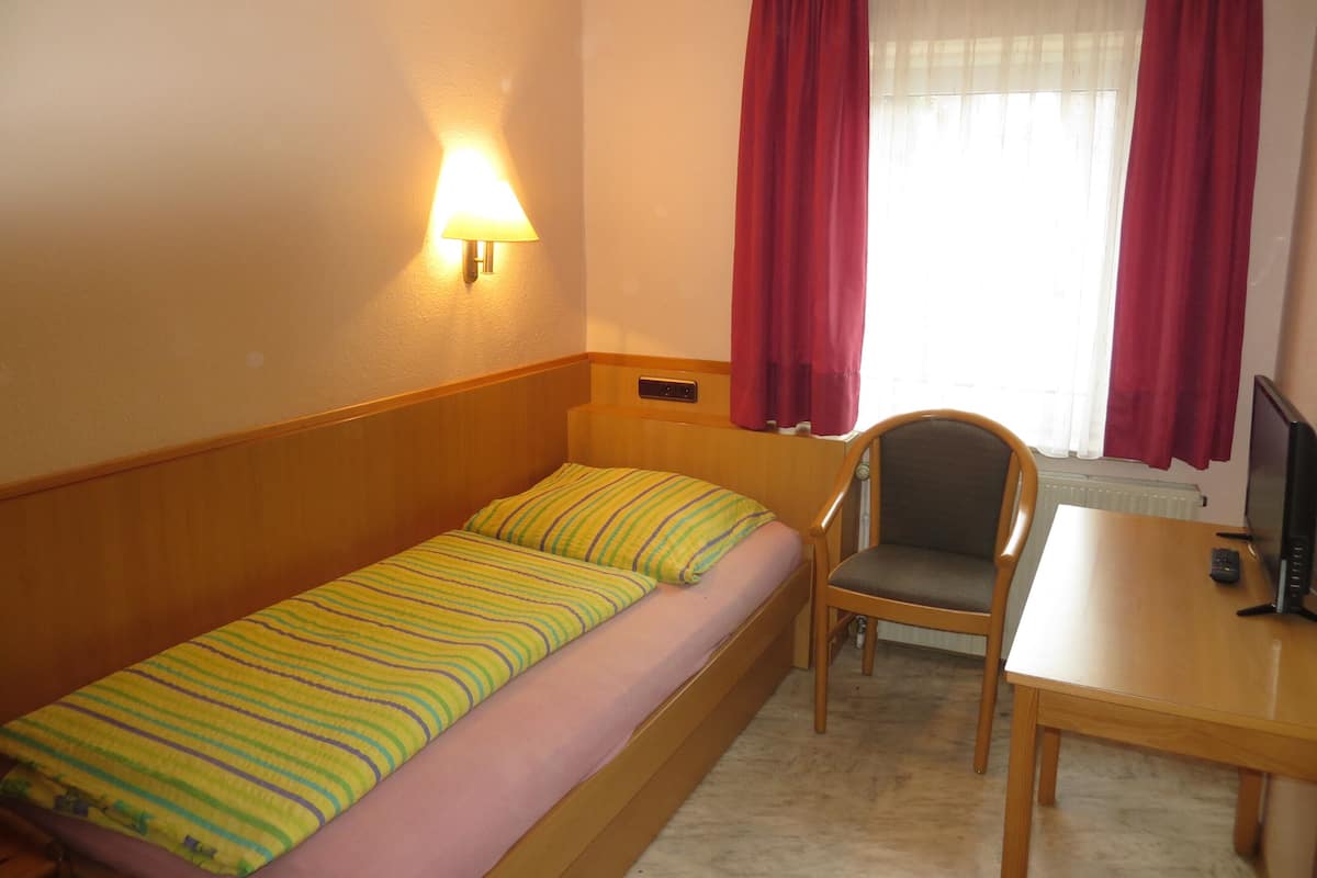 A small hotel room with a single bed, a desk, a chair, a TV, and a window with red curtains.