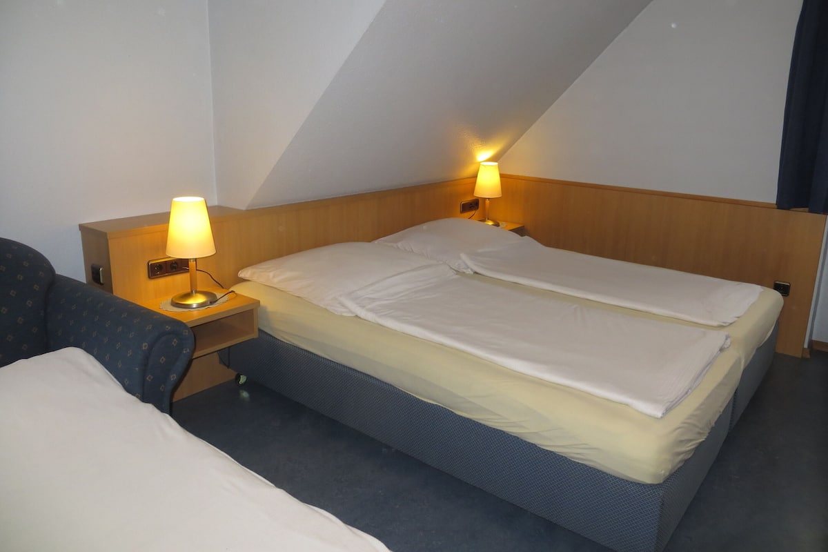 A hotel room with a single bed, two bedside lamps, a wooden headboard, and a small bedside table.