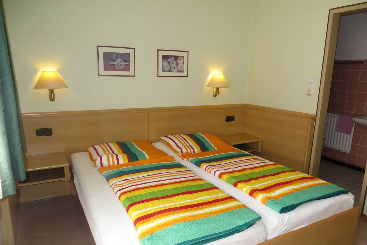 A double bed with a striped blanket, two wall-mounted lamps, and two framed pictures on the wall.
