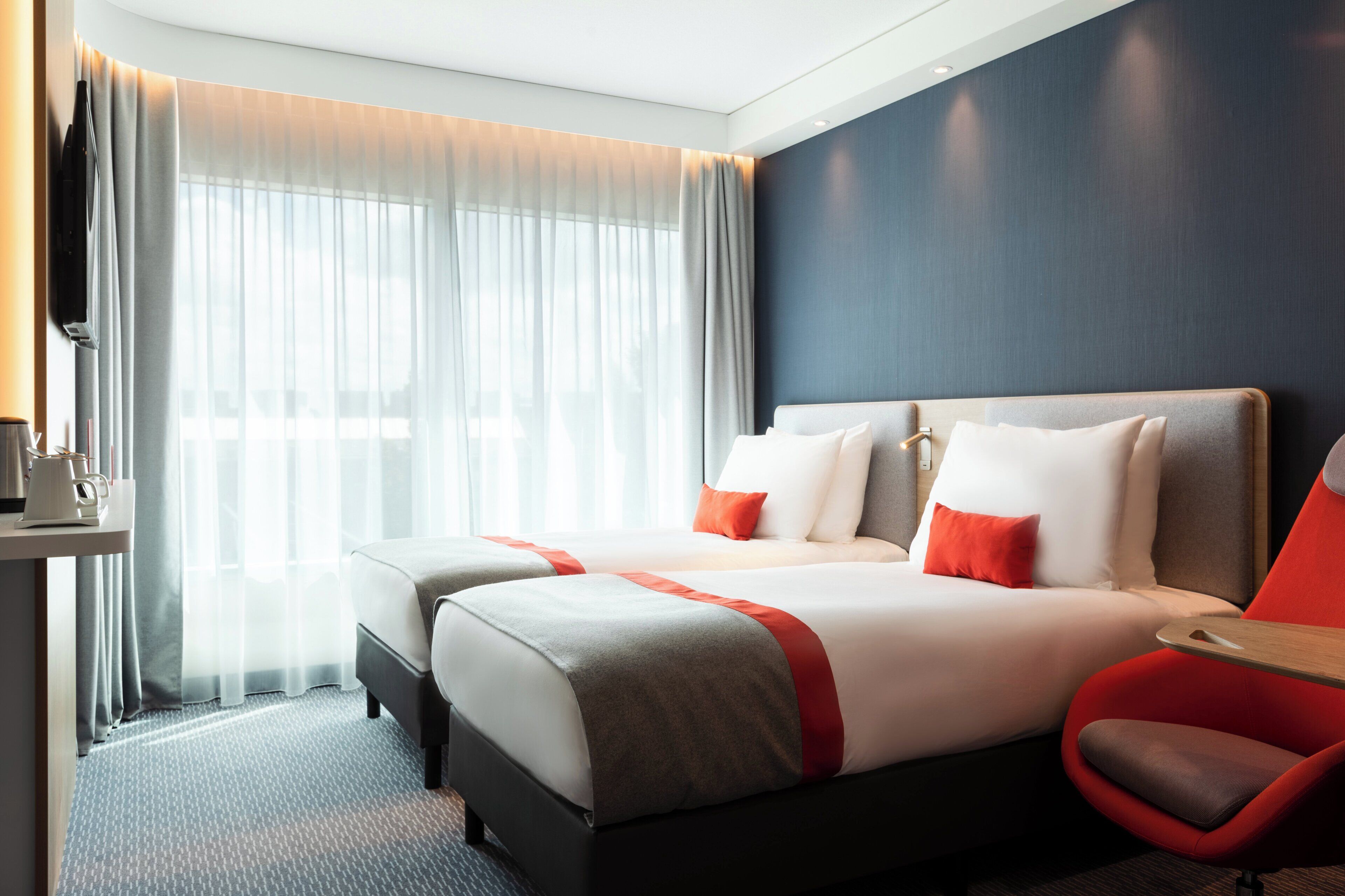 A hotel room with two beds, a TV, a desk with a coffee maker, and a red chair.