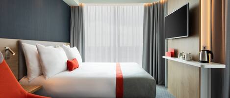 Habitación de hotel moderna con una cama grande, un televisor montado en la pared, una mesita de noche con hervidor y vistas a la ciudad a través de cortinas translúcidas.