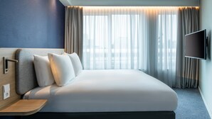 A modern hotel room with a large bed, a flat-screen TV mounted on the wall, and a city view through sheer curtains. - Holiday Inn Express Almere by IHG (Almere)