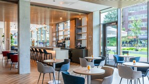 A modern cafe with round tables and chairs, a bar area, and large windows offering a view of the street. - Holiday Inn Express Almere by IHG (Almere)