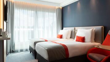 A hotel room with two beds, a TV, a desk with a coffee maker, and a large window with sheer curtains.