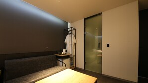 A modern hotel room with a wooden table, a gray sofa, a small shelf with a white robe and a coffee maker, and a sliding glass door leading to a bathroom. - Hotel Yeoubi (Busan)