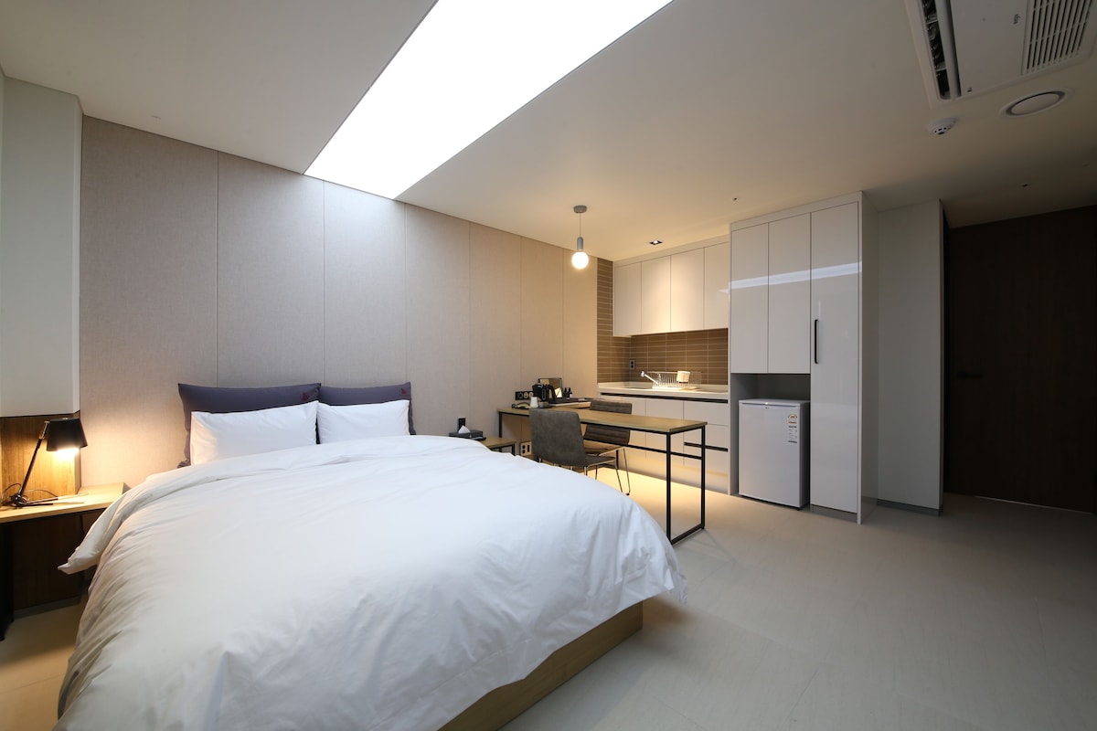 A modern hotel room with a large bed, a desk, and a kitchenette.