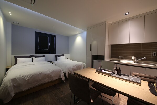 A modern hotel room with two beds, a wooden desk, a small kitchenette, and a window with a view of the city.