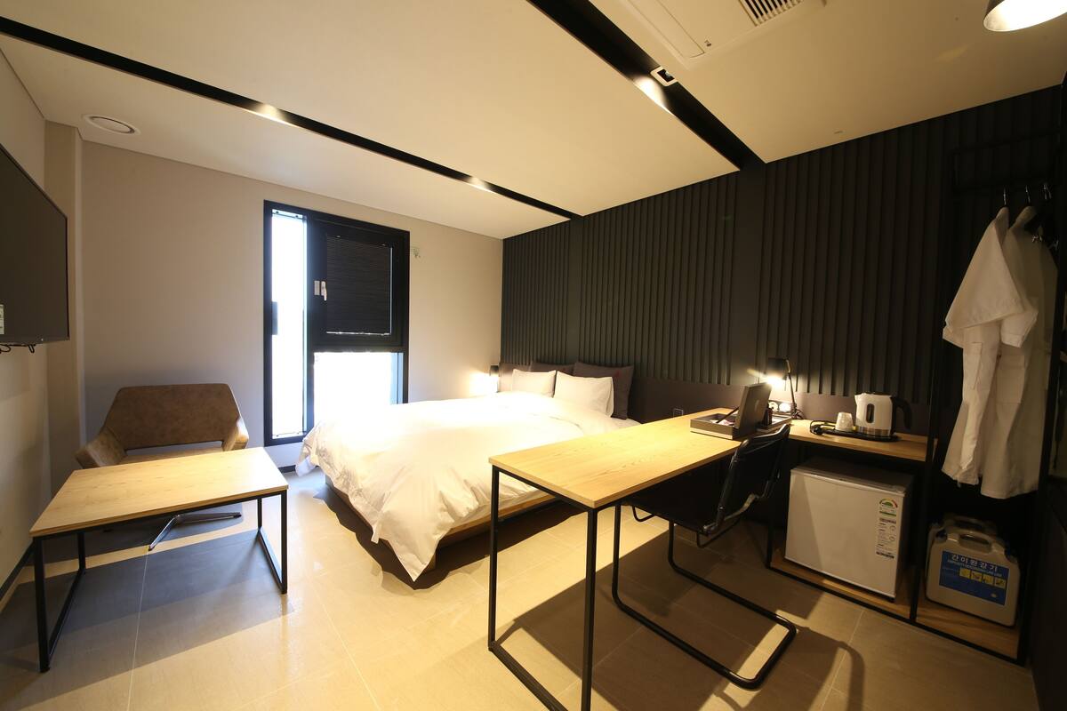 Una habitación de hotel moderna con cama, escritorio, silla, refrigerador pequeño y ventana con persianas.