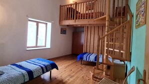 Ein Zimmer mit Holzboden, zwei Einzelbetten mit blau-weißer Bettwäsche, einem Holzstuhl, einer Holztreppe und einem Fenster mit Jalousien.