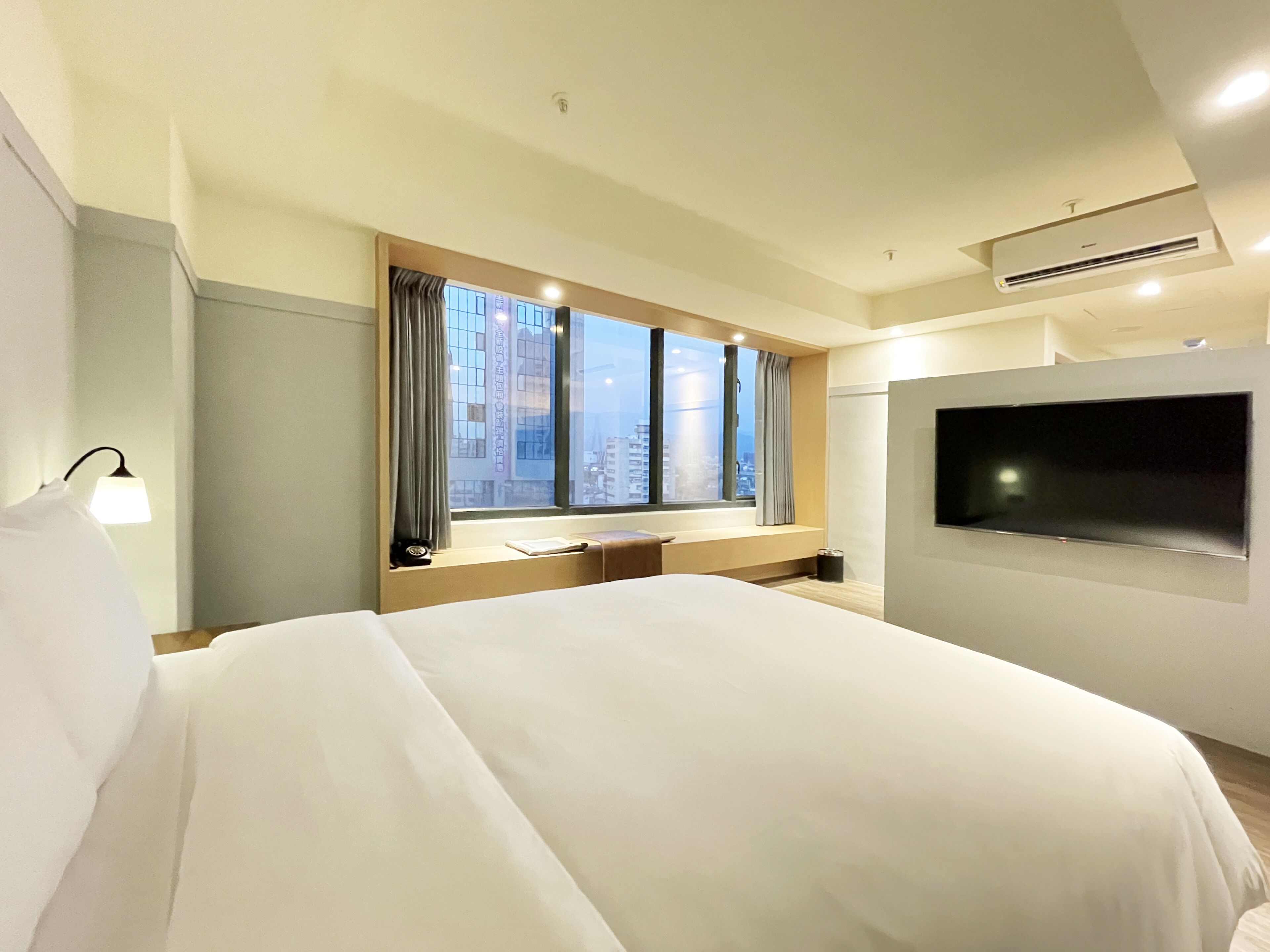Ein Hotelzimmer mit einem großen Bett, einem an der Wand befestigten Fernseher, einem Fenster mit Vorhängen und Blick auf die Stadt.