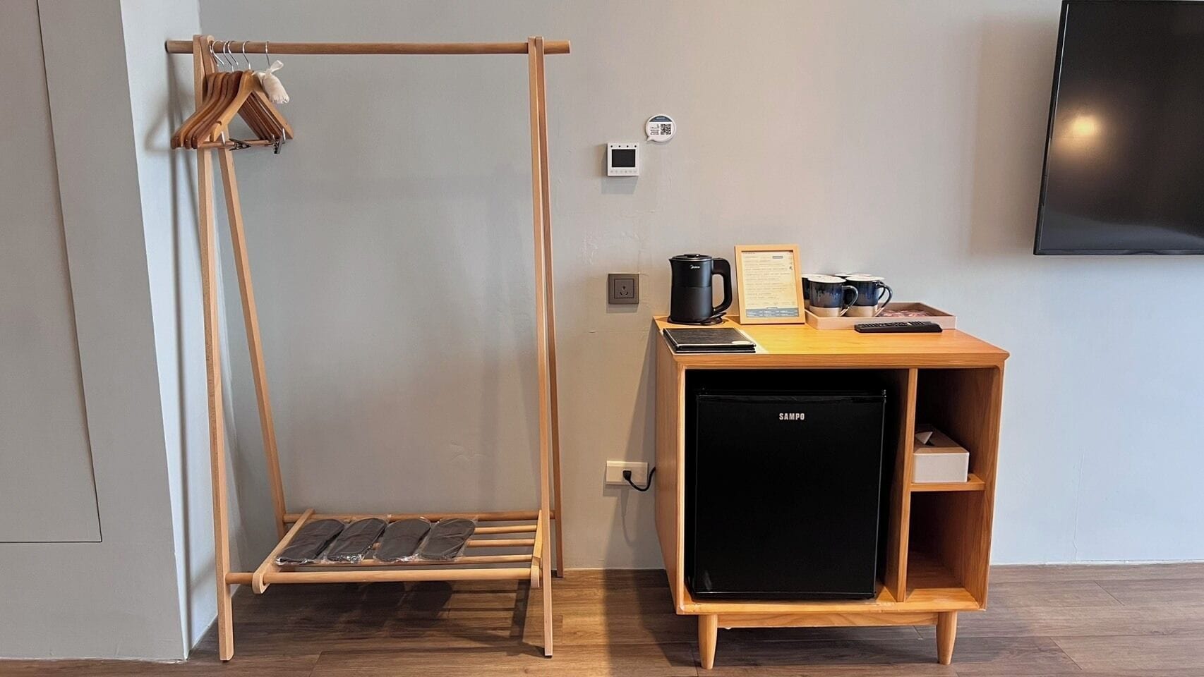 Une pièce comprenant un bureau en bois, un réfrigérateur noir, un porte-manteau en bois et un téléviseur fixé au mur.
