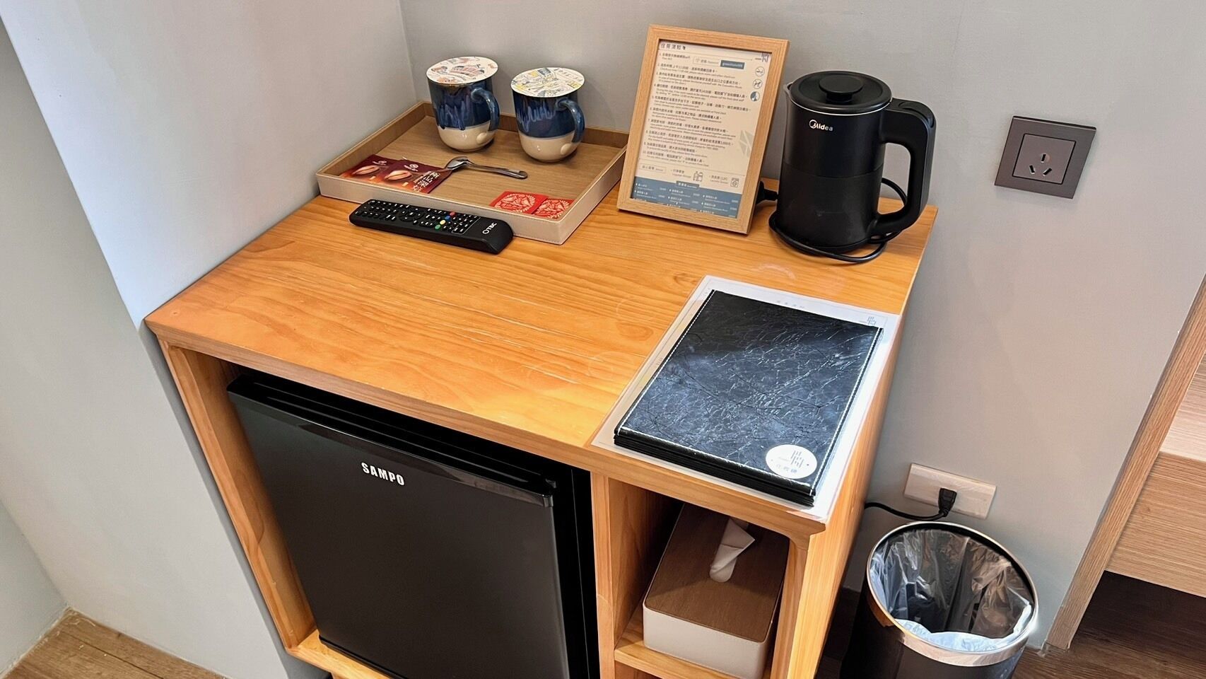 A compact kitchenette with a black refrigerator, a coffee maker, a tray with two mugs, a remote control, and a menu board.