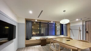 A modern hotel room with a large window, a dining area with a wooden table and chairs, and a flat-screen TV mounted on the wall. - Green Hotel - midori (Taichung)