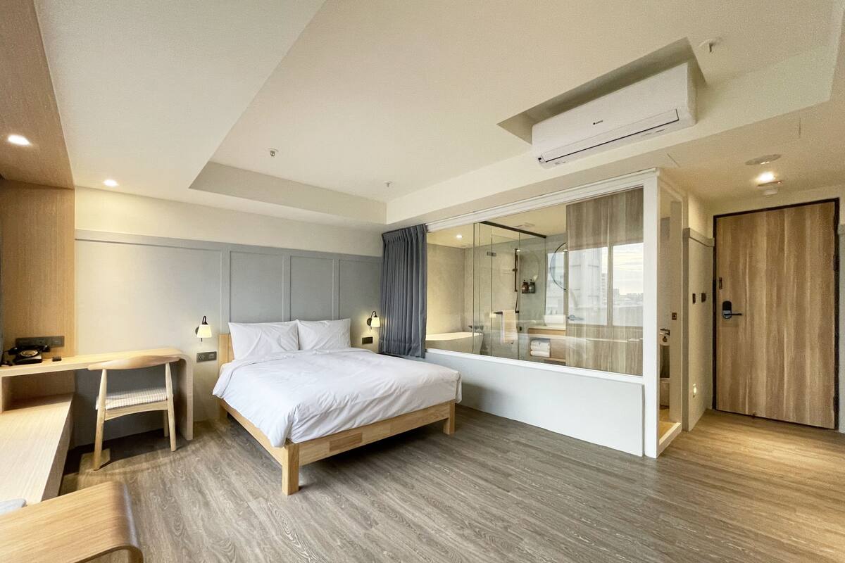 A modern hotel room with a wooden floor, a bed with white linens, a desk, a chair, a shower area, and a large mirror.