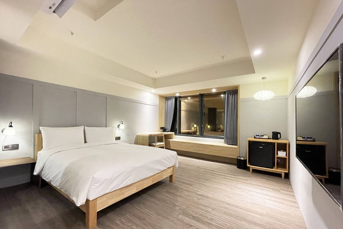A modern hotel room with a large bed, bedside tables, a desk, a mirror, and a wardrobe.