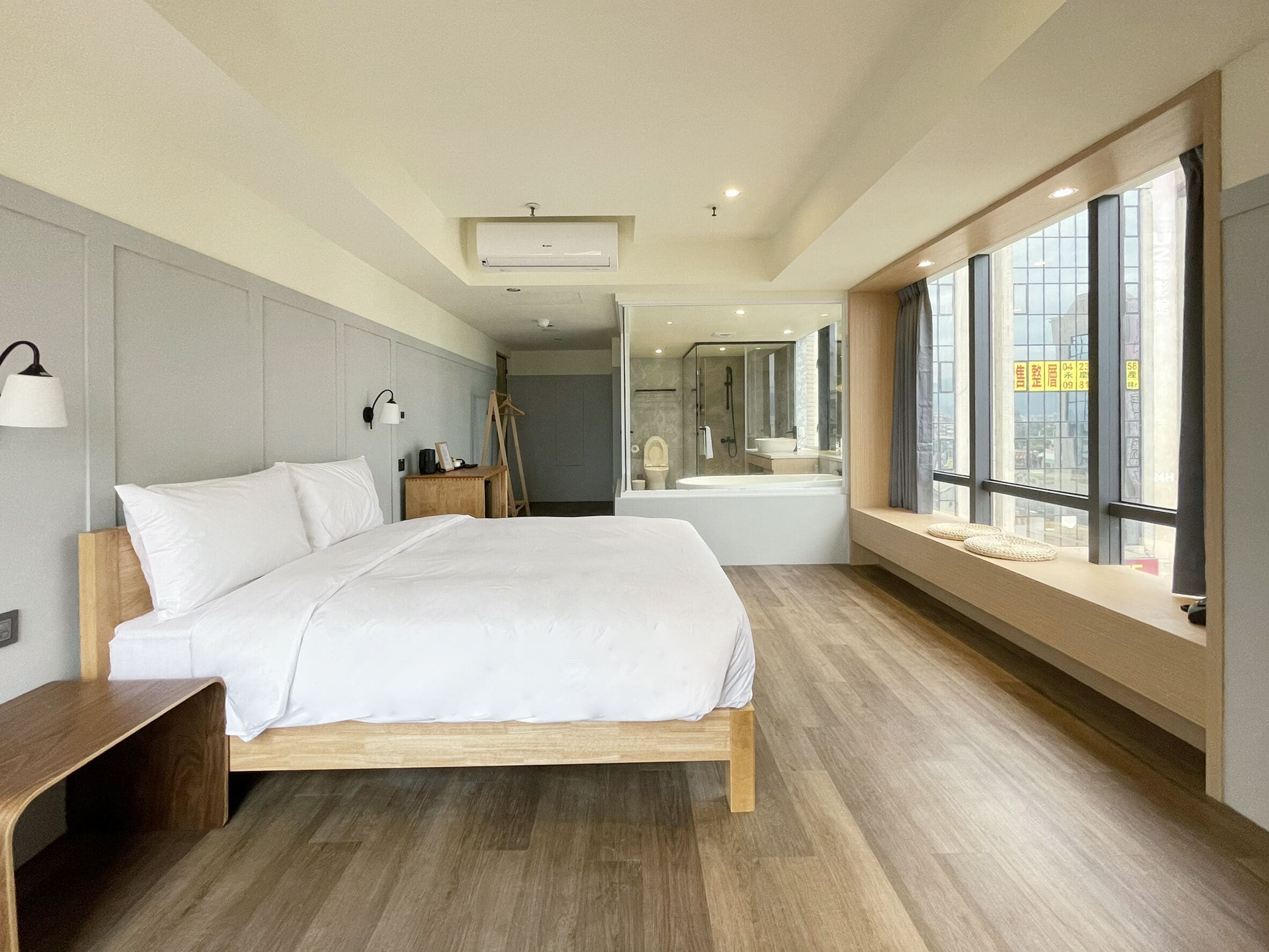 A modern hotel room with a large bed, a wooden bedside table, a bedside lamp, a window with a view, and a bathroom with a bathtub and shower.