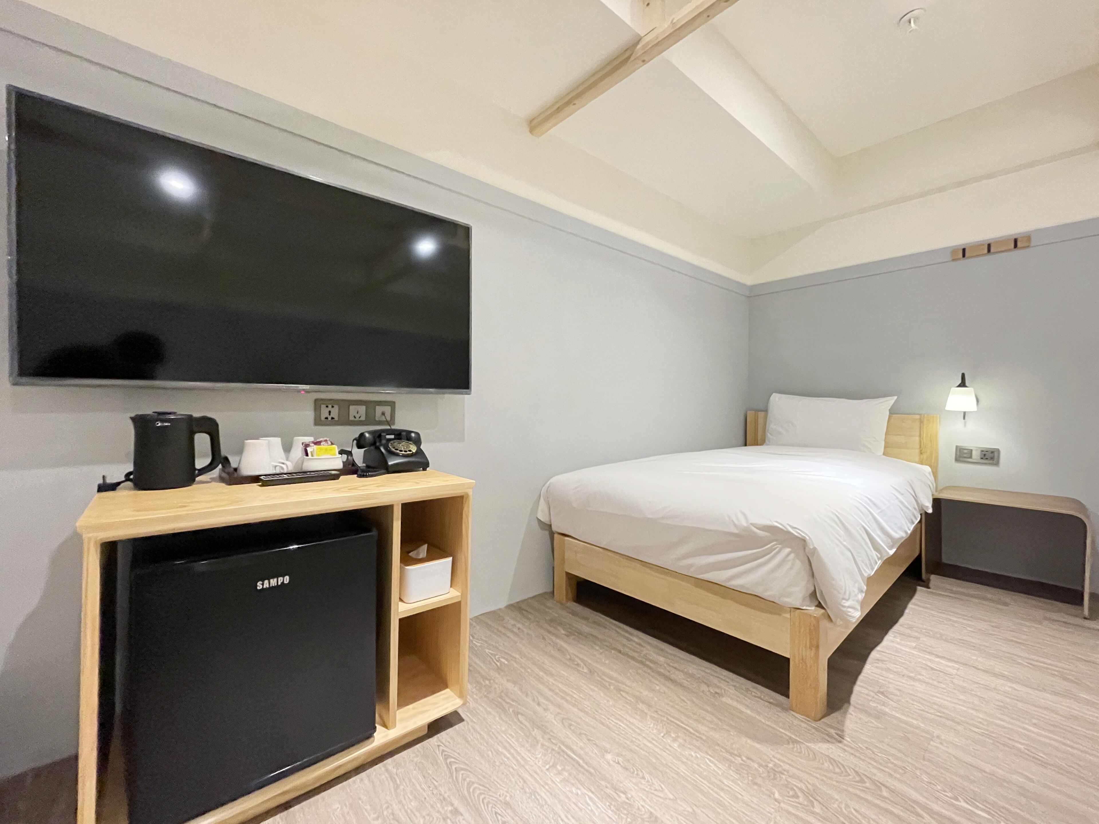 A hotel room with a bed, a TV mounted on the wall, a small desk with a kettle, a refrigerator, and a bedside lamp.