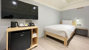 A hotel room with a bed, a TV mounted on the wall, a small desk with a kettle, a refrigerator, and a bedside lamp.