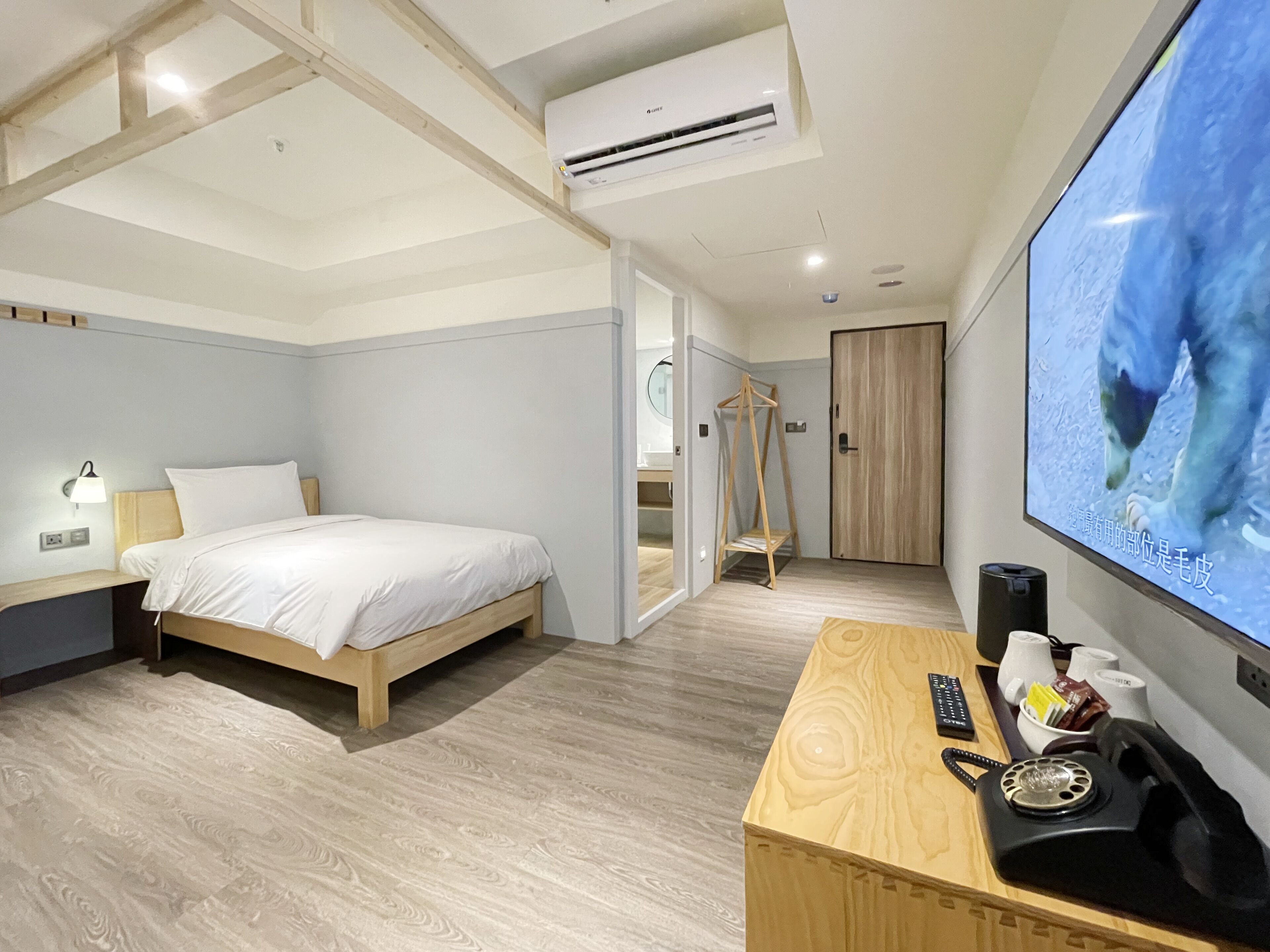 Quarto de hotel com cama, mesa de cabeceira, televisão e escrivaninha de madeira com telefone.