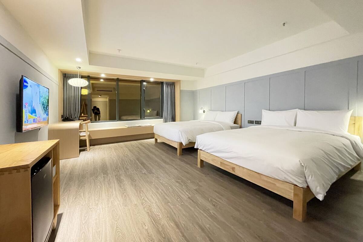 A hotel room with two beds, a TV, a desk, and a large window.