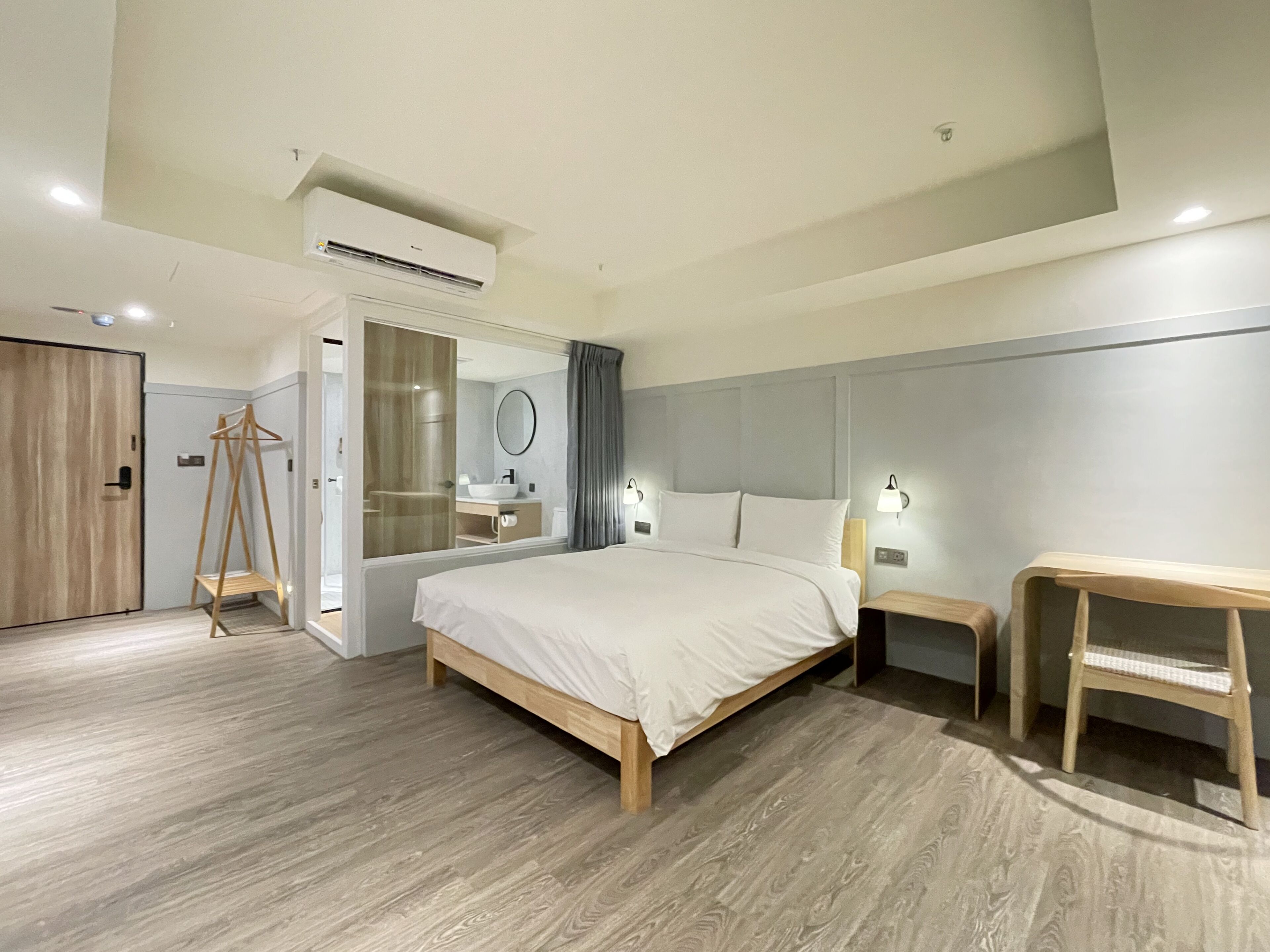 A modern hotel room with a large bed, a wooden floor, a bathroom with a mirror, and a wardrobe.