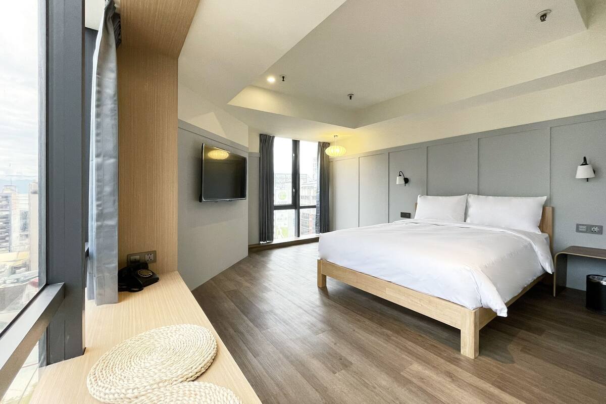 A modern hotel room with a large bed, a flat-screen TV mounted on the wall, and a large window offering a city view.