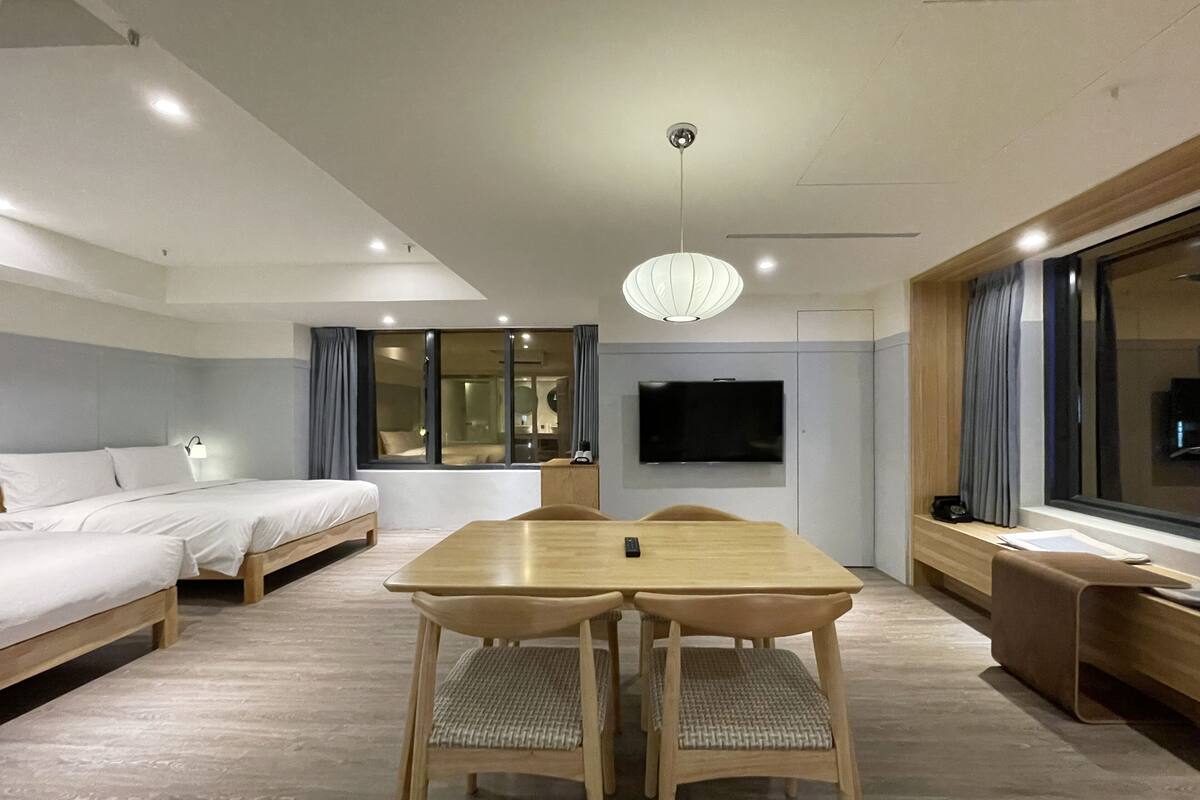 A modern hotel room with a wooden dining table, two chairs, a sofa, a TV mounted on the wall, and a large window with curtains.