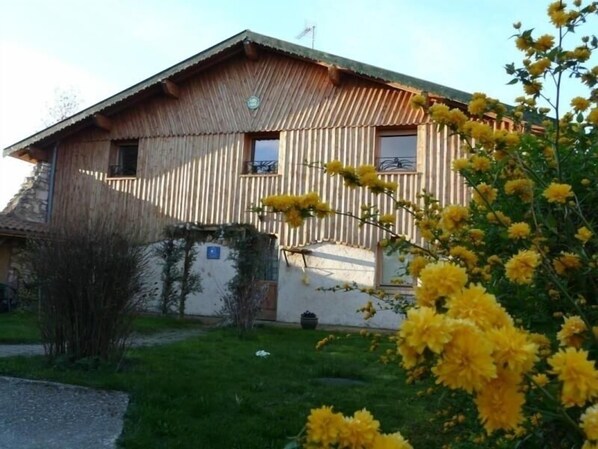 A wooden house with a sloped roof, a small balcony, and a garden with yellow flowers.