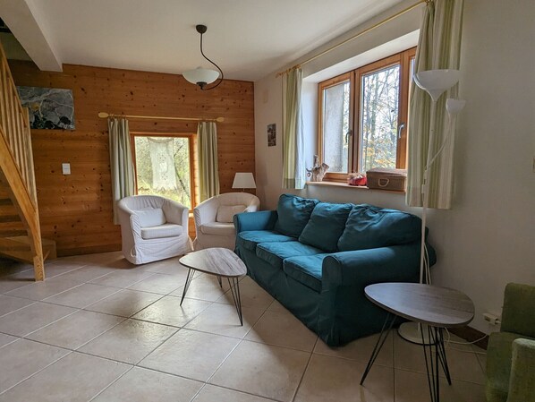 A cozy living room with a wooden floor, a blue sofa, two white armchairs, a small round table, and a staircase leading to another level. - Riverside Mill: Renovated Historic Retreat in Lorraine Nature Park (Bonzée)