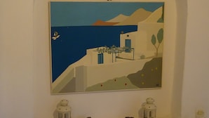 Una pintura de una escena costera con un edificio blanco, una mesa con una puerta azul y dos faroles en el suelo.