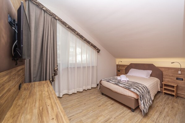 Una camera da letto con un letto, un comodino in legno, una lampada da comodino, una TV fissata al muro e una finestra con tende.