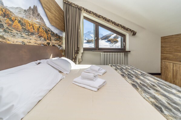 Une chambre d’hôtel avec un lit, une fenêtre donnant sur la montagne, un radiateur et une tête de lit en bois.