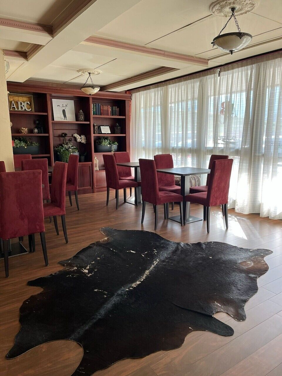 A room with a wooden floor, red chairs, a black leather rug, a bookshelf, and a large window with sheer curtains.