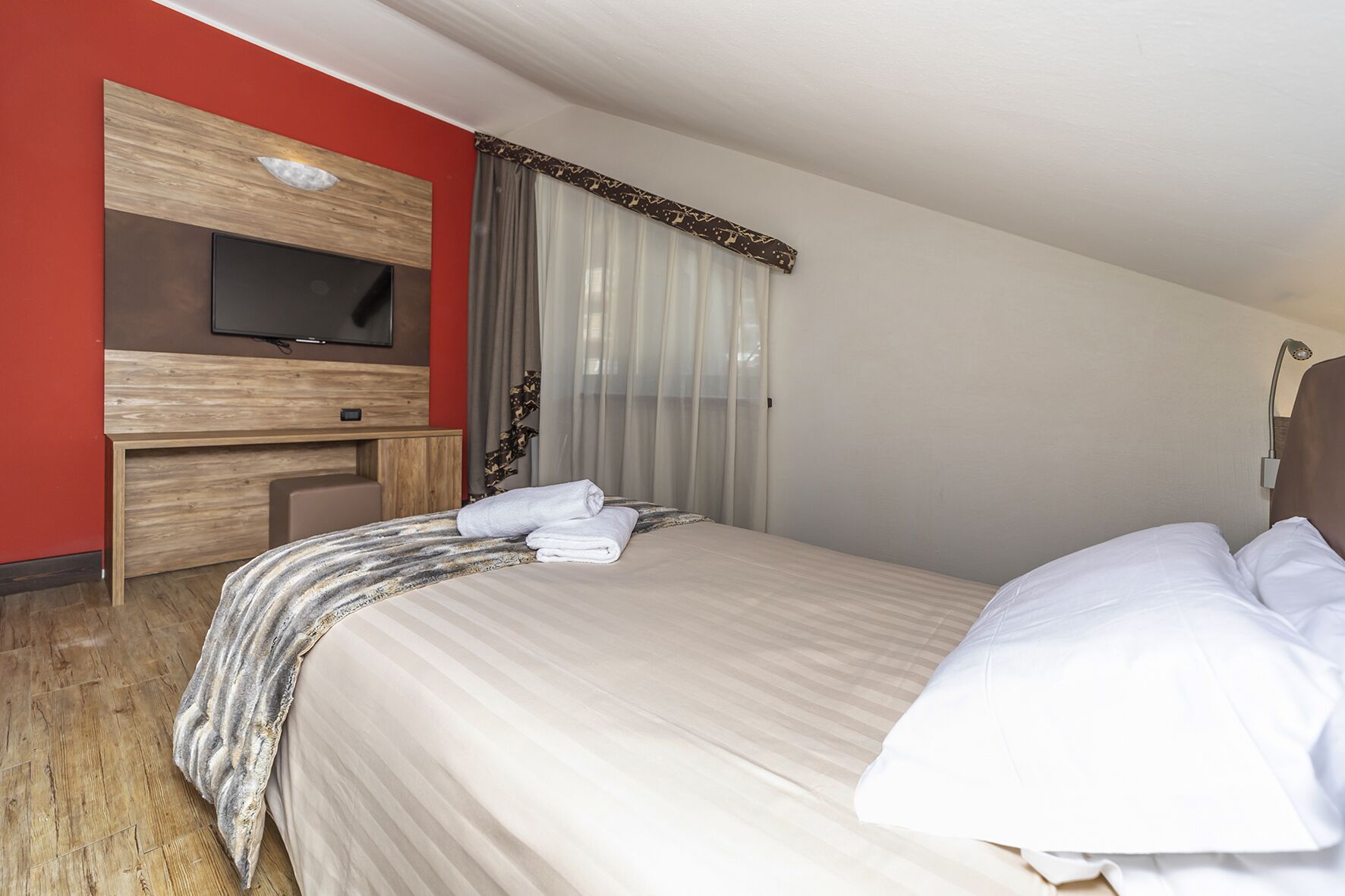 Una habitación de hotel con cama, televisor montado en un panel de madera, escritorio y ventana con cortinas traslúcidas.