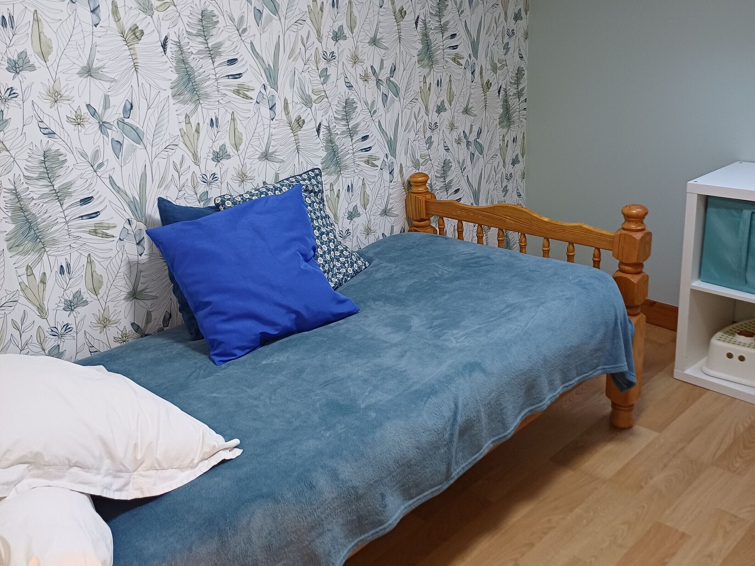 A bedroom with a bed, pillows, a wooden headboard, a wall with a leaf pattern, and a white shelf unit.