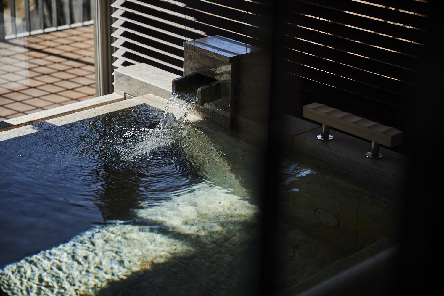 A water feature with a stone basin and a small waterfall, located indoors with a window and blinds in the background.