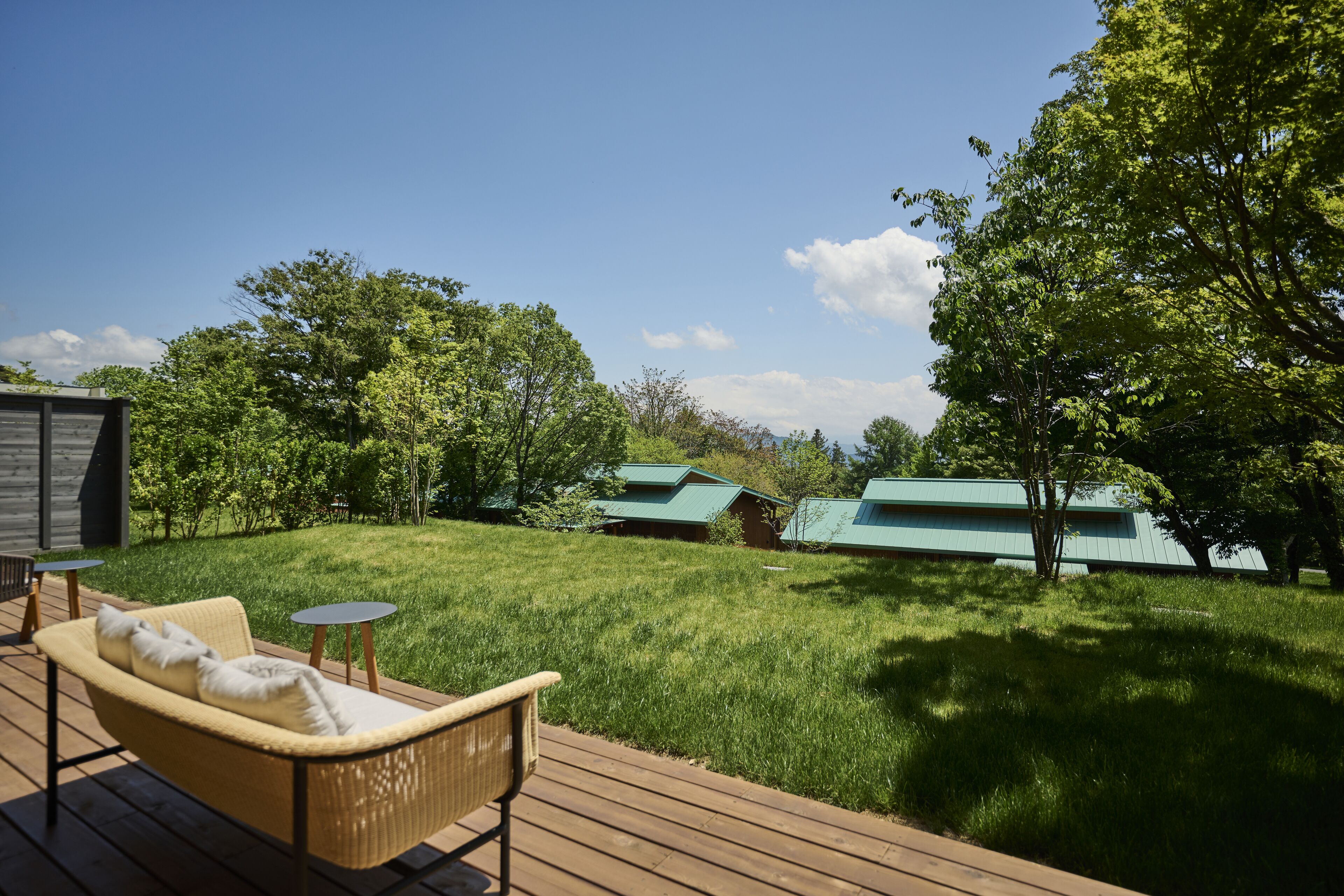 A wooden deck with a wicker sofa and two small tables, a grassy lawn, and several buildings with green roofs in the background.