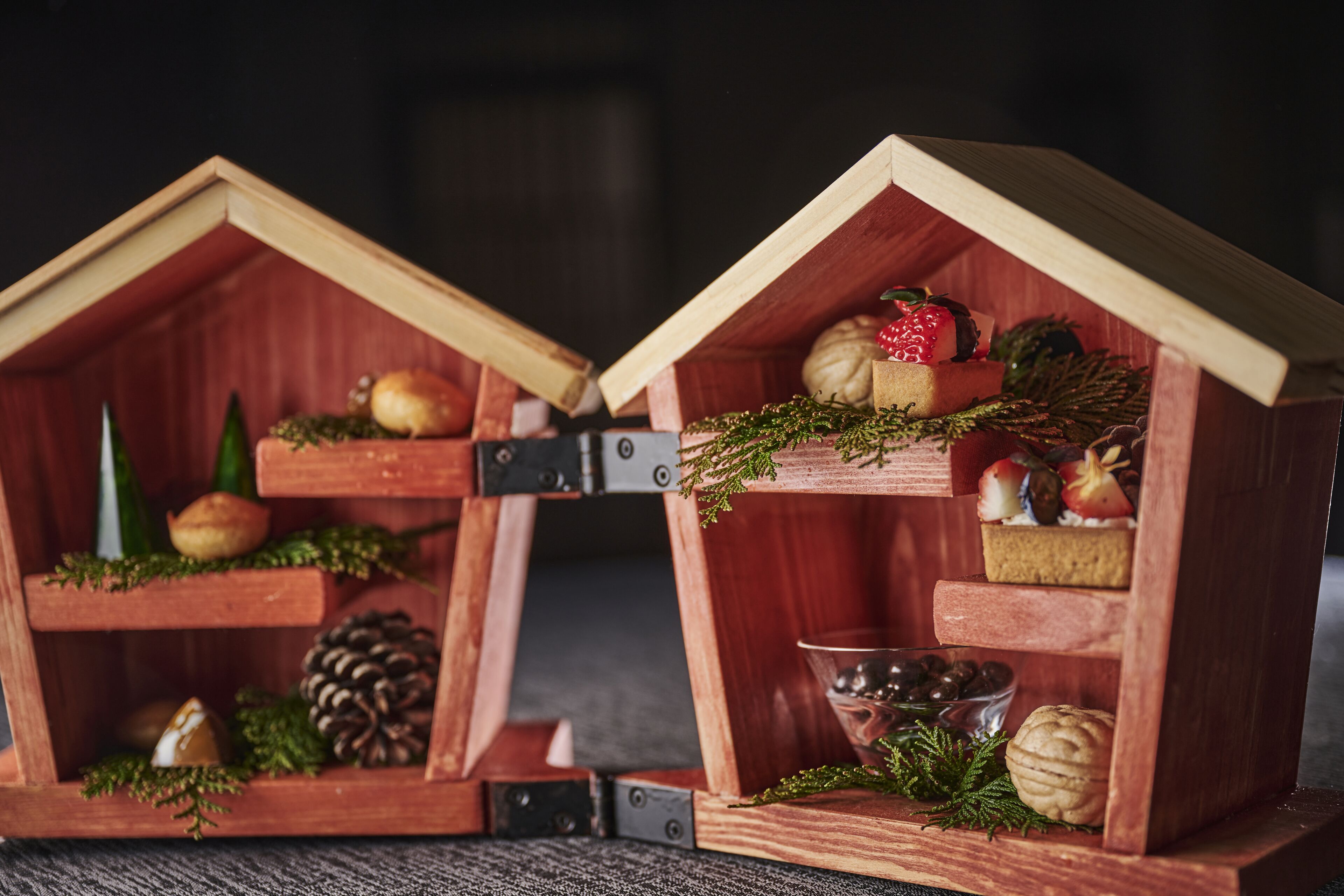 Two wooden miniature houses with shelves containing various items like pine cones, a pine tree, and a bowl.