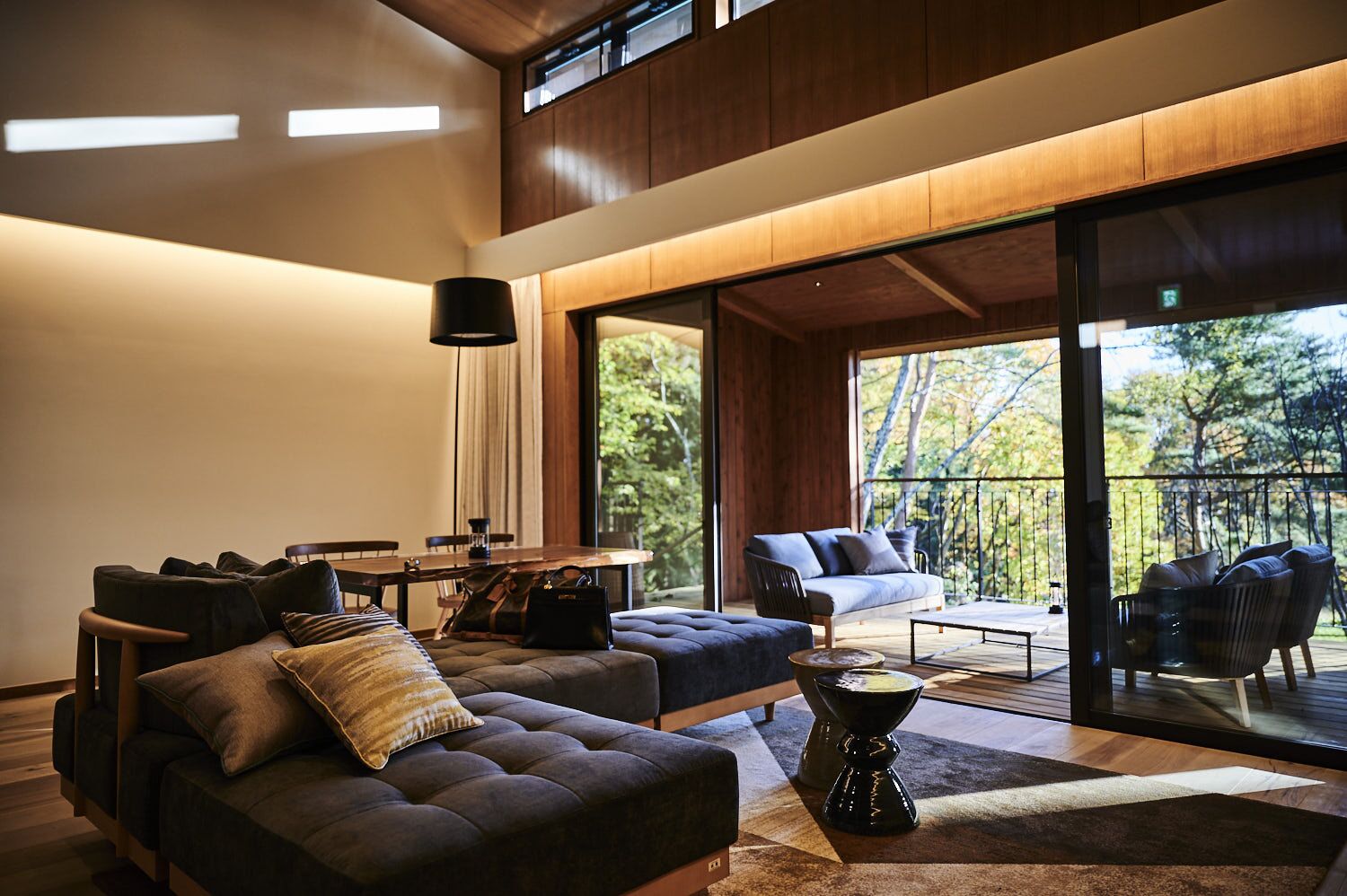 A modern living room with a sofa, armchair, and dining table. There is a balcony with outdoor furniture.