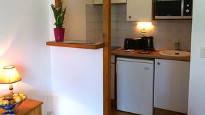Eine kompakte Küche mit Mikrowelle, Toaster und kleinem Kühlschrank. Auf einem Regal steht eine Topfpflanze, und ein Obstschälchen steht auf einem Beistelltisch.