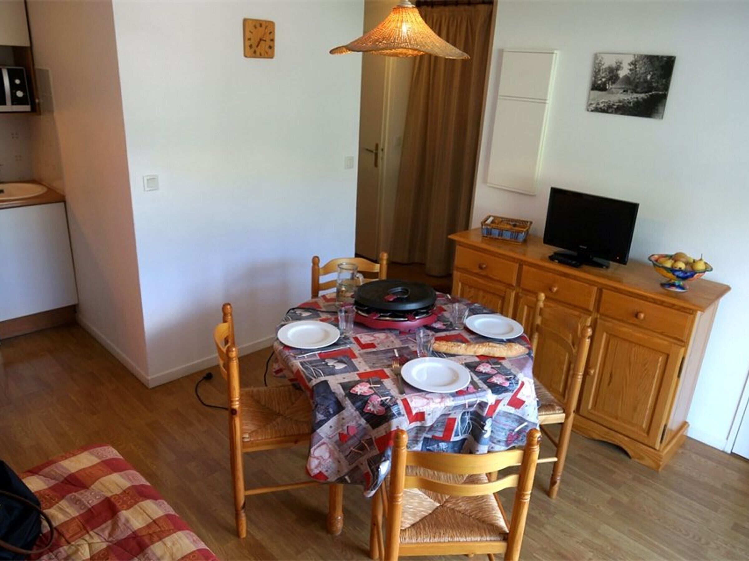Un espacio habitable compacto con zona de comedor, una mesa dispuesta para dos personas, un televisor sobre un mueble de madera, y una pequeña kitchenette al fondo.