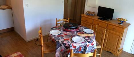 Un espacio habitable compacto con zona de comedor, una mesa dispuesta para dos personas, un televisor sobre un mueble de madera, y una pequeña kitchenette al fondo.