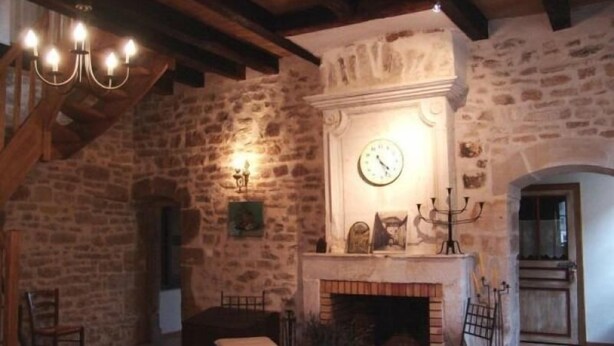 A rustic room with exposed stone walls, a wooden ceiling, a fireplace with a clock above it, and a chandelier. - Historic XIV Century Cottage with Modern Comforts, Garden and Parking (Void-Vacon)