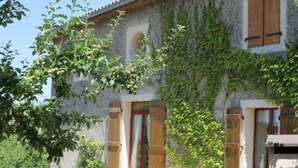 Un edificio de piedra con contraventanas de madera, un pequeño jardín y un árbol en primer plano.