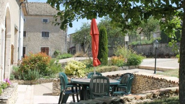 Un edificio de piedra con un patio que incluye una mesa y sillas, rodeado de vegetación y un muro de piedra.