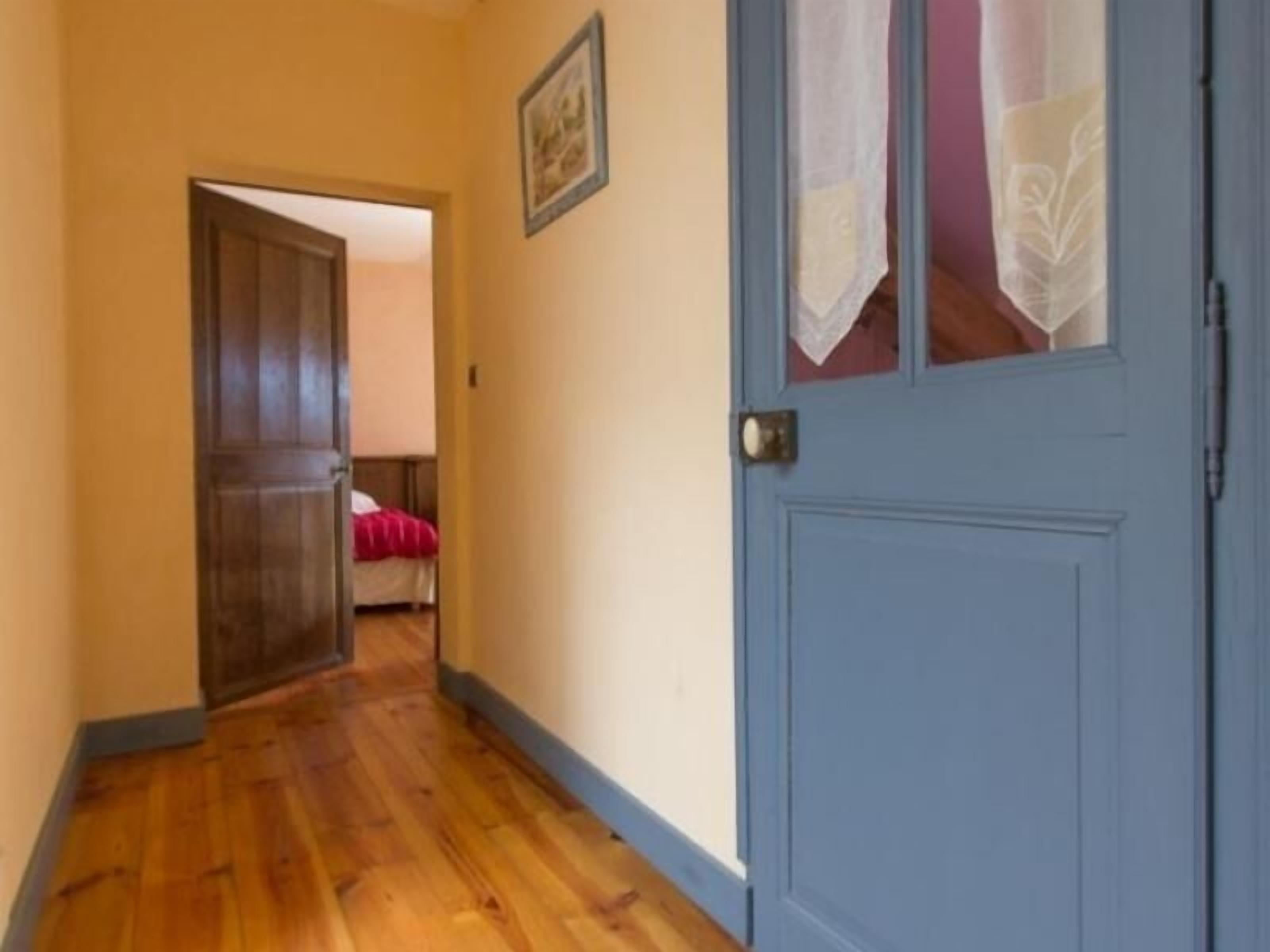 A hallway with wooden flooring, a blue door, and a bed with a red blanket in the background.