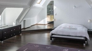 Une chambre moderne et bien éclairée, située sous les combles, avec une fenêtre de toit, un lit, une commode et une vue sur la salle de bain.