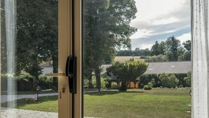 Une vue à travers une porte vitrée dotée d’une poignée en métal, donnant sur une cour verdoyante avec des arbres et un bâtiment dont le toit est recouvert de tuiles.