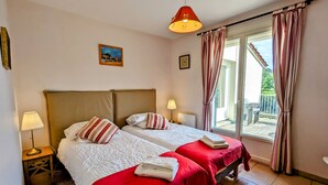Ein Doppelzimmer mit zwei Betten, jeweils mit roter Tagesdecke und weißen Kissen, einem kleinen Nachttisch mit Lampe, einem Bild an der Wand und Blick durch eine Schiebetür nach draußen.